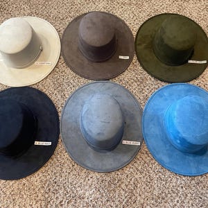 May include: Six suede-like hats in various colors are displayed. Colors include light gray, gray, olive green, navy blue, blue jeans, and turquoise. Each hat has a wide brim and a cylindrical crown. Small labels identify each color.