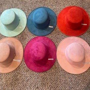 May include: Six colorful hats in shades of mint, teal, red, fuchsia, rosewood, and pink baby. Each hat has a wide brim and a cylindrical crown. The hats are arranged on a textured surface.