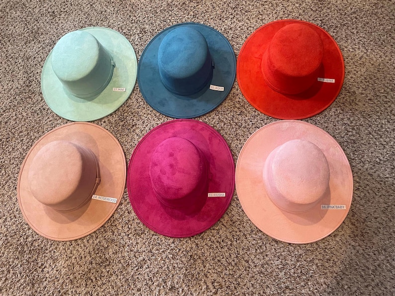 May include: Six colorful hats in shades of mint, teal, red, fuchsia, rosewood, and pink baby. Each hat has a wide brim and a cylindrical crown. The hats are arranged on a textured surface.