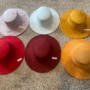 May include: Six suede-like hats in various colors are displayed on a carpeted surface. The hats include lavender, light blue, mustard, red, wine, and pumpkin. Each hat has a tag with a color name. The hats have a classic fedora style.