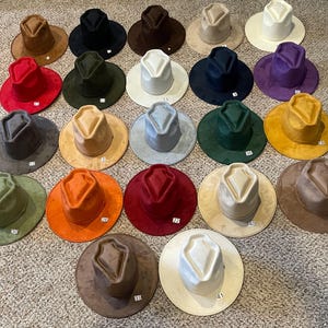 Puede incluir: Una colección de 21 sombreros en varios colores y materiales. Los sombreros son todos de estilo fedora con un ala ancha y una corona en forma de diamante. Los sombreros están dispuestos en semicírculo sobre una superficie marrón claro.