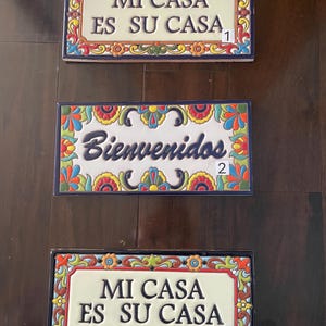 Könnte beinhalten: Drei farbige Keramikfliesen mit spanischen Sätzen. Die obere Fliese lautet "MI CASA ES SU CASA" mit der Nummer 1. Die mittlere Fliese lautet "Bienvenidos" mit der Nummer 2. Die untere Fliese lautet "MI CASA ES SU CASA" mit der Nummer 3.