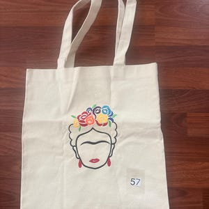 May include: A cream-colored canvas tote bag featuring a black line-art illustration of a face with a floral crown in red, yellow, blue, and green. The face has red earrings and red lips. A small white square label with the number 57 is attached.