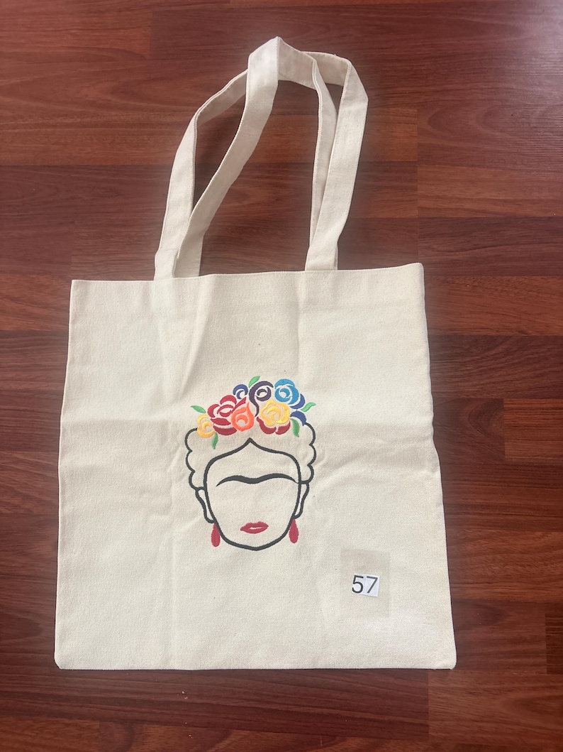 May include: A cream-colored canvas tote bag featuring a black line-art illustration of a face with a floral crown in red, yellow, blue, and green. The face has red earrings and red lips. A small white square label with the number 57 is attached.