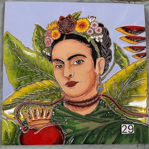 May include: A square ceramic tile featuring a portrait of Frida Kahlo. She wears a floral crown, earrings, and a necklace. The image includes large green leaves, a red heart with a crown, and the number 29.