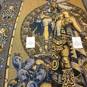 May include: A woven quilt with a detailed Aztec-inspired design. The quilt features a central image of a figure with a feathered headdress, in shades of gold, blue, and black. The border includes geometric patterns and cactus motifs. The text on the label reads "Queen Size".