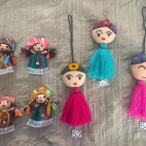 May include: A collection of seven handmade doll keychains featuring embroidered faces, colorful fabric dresses with lace trim, and yarn tassels. The dolls are labeled with small white tags numbered 5, 7, 24, 33, 52, 53, and 54, each with unique hair and headpiece details.