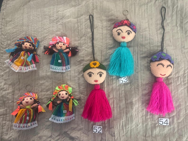 May include: A collection of seven handmade doll keychains featuring embroidered faces, colorful fabric dresses with lace trim, and yarn tassels. The dolls are labeled with small white tags numbered 5, 7, 24, 33, 52, 53, and 54, each with unique hair and headpiece details.