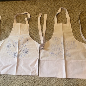May include: Two white aprons with blue embroidered designs. The apron on the left has a floral design with a bird in the centre. The apron on the right has a bird design with flowers around it. The aprons are numbered 6 and 8.