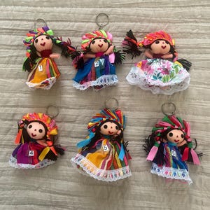 May include: Six handmade doll keychains featuring fabric bodies with black hair, colorful ribbon headpieces, and dresses with lace trim. Each doll is numbered one through six on the front of the skirt, and includes a metal ring for attachment.