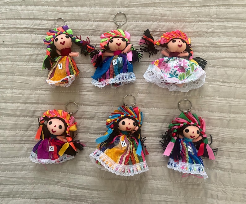 May include: Six handmade doll keychains featuring fabric bodies with black hair, colorful ribbon headpieces, and dresses with lace trim. Each doll is numbered one through six on the front of the skirt, and includes a metal ring for attachment.
