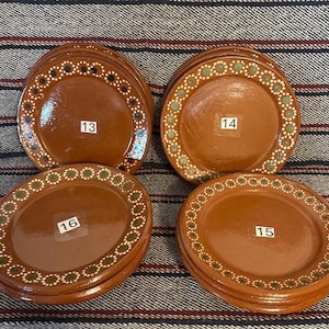 Op de afbeelding: Vier stapels ronde bruine keramische borden met een glanzende afwerking. Elk bord heeft een decoratieve rand van kleine witte en groene stippen. Witte rechthoekige labels met de nummers 13, 14, 15 en 16 zijn in het midden van elke stapel bevestigd.