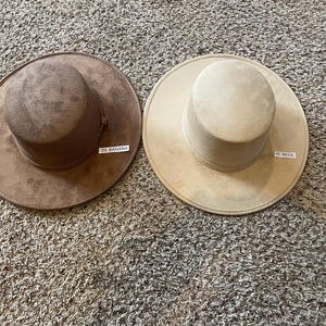 May include: Two suede-like hats in the colors of banana and beige. The hat on the left is brown with a tag that reads "39. BANANA". The hat on the right is beige with a tag that reads "40. BEIGE". Both hats have a wide brim and a cylindrical crown.