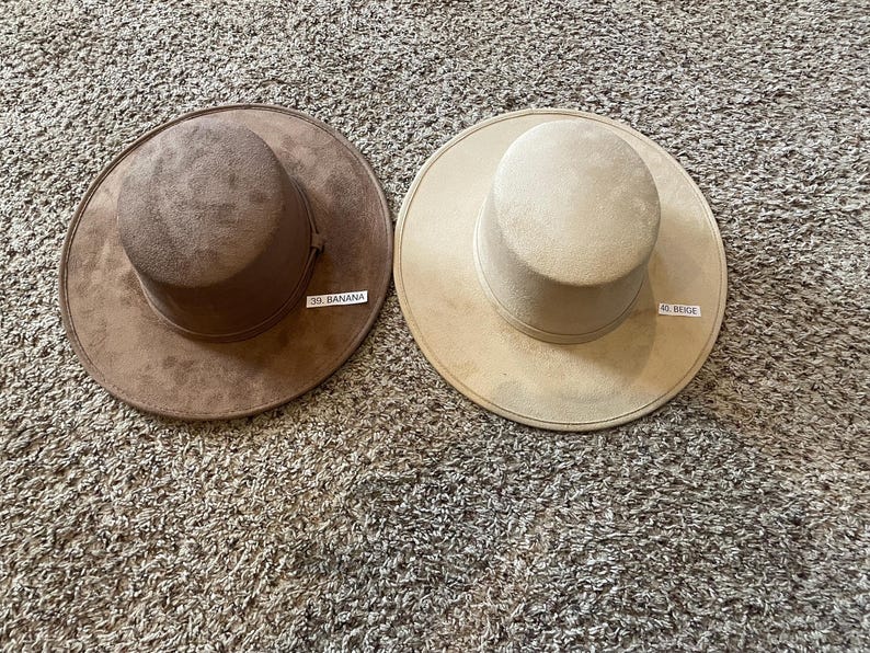 May include: Two suede-like hats in the colors of banana and beige. The hat on the left is brown with a tag that reads "39. BANANA". The hat on the right is beige with a tag that reads "40. BEIGE". Both hats have a wide brim and a cylindrical crown.