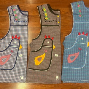 May include: Three sleeveless aprons featuring a checkered pattern in blue, brown, and light blue. Each apron includes a large chicken-shaped pocket with a red comb, yellow or pink wing, and embroidered details. Small numbered tags 1, 2, and 3 are visible.