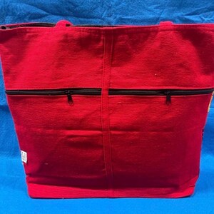 May include: A bright red tote bag featuring a textured fabric surface and two black horizontal zippered pockets on the front. The bag includes two top handles and a small white rectangular tag attached to the lower left side of the exterior.