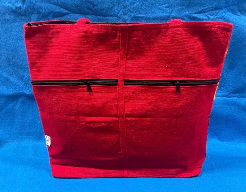 May include: A bright red tote bag featuring a textured fabric surface and two black horizontal zippered pockets on the front. The bag includes two top handles and a small white rectangular tag attached to the lower left side of the exterior.