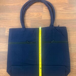 May include: A navy blue tote bag with two top handles and a horizontal zippered pocket on the front. A yellow measuring tape is placed vertically over the center, showing the bag height is approximately 14 inches from the base to the top edge.