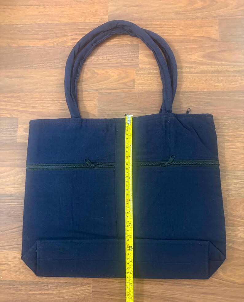 May include: A navy blue tote bag with two top handles and a horizontal zippered pocket on the front. A yellow measuring tape is placed vertically over the center, showing the bag height is approximately 14 inches from the base to the top edge.
