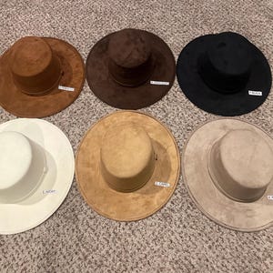May include: Six suede-like hats in various colors: tan, brown, black, ivory, camel, and mocha. Each hat has a wide brim and a cylindrical crown. The hats are labeled with color names.