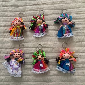 May include: Six fabric doll keychains with black yarn hair, colorful ribbon headpieces, and dresses featuring lace trim. Each doll has a small white tag numbered 7, 8, 9, 10, 11, or 12 attached to the front. The dolls feature various patterns.