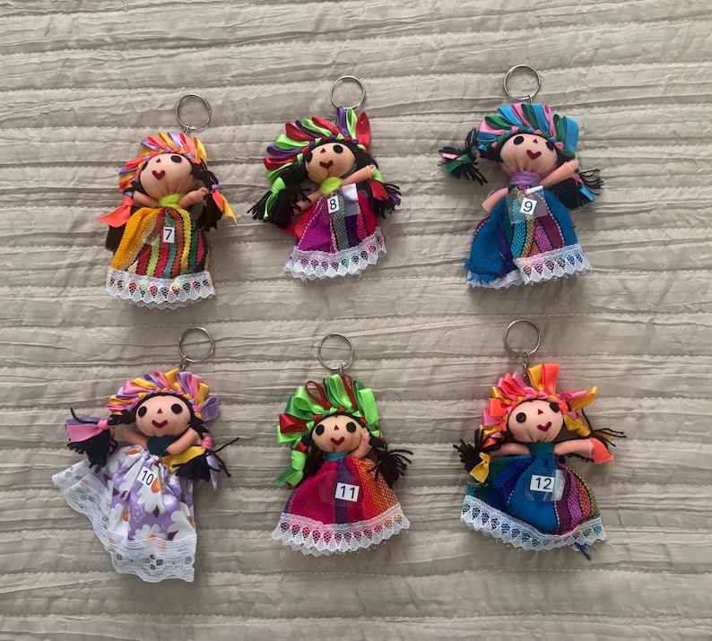 May include: Six fabric doll keychains with black yarn hair, colorful ribbon headpieces, and dresses featuring lace trim. Each doll has a small white tag numbered 7, 8, 9, 10, 11, or 12 attached to the front. The dolls feature various patterns.