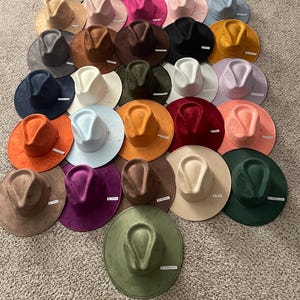 Könnte beinhalten: Eine Sammlung von einunddreißig Fedora-Hüten mit breiter Krempe, die in einem Raster angeordnet sind und eine vielfältige Auswahl an einfarbigen Modellen in Rot, Blau, Grün, Gelb, Orange, Lila, Pink, Schwarz, Weiß, Grau, Beige und Braun zeigen.