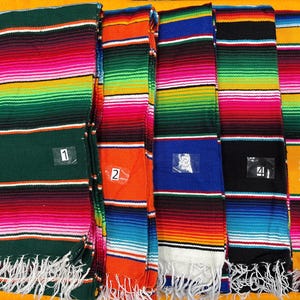 May include: A collection of colorful serape blankets with horizontal stripes in various colors, including green, pink, orange, blue, black, and yellow. Each blanket has a white fringe at the bottom. Numbers 1 through 5 are visible on the blankets.