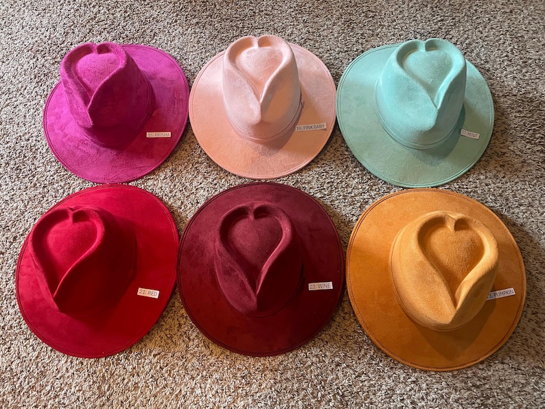 K&ouml;nnte beinhalten: Sechs Filzh&uuml;te in verschiedenen Farben mit einem herzf&ouml;rmigen Design auf der Krone. Die H&uuml;te sind in einem Kreis auf einer strukturierten Oberfl&auml;che angeordnet. Die Farben der H&uuml;te sind rosa, hellrosa, hellblau, rot, burgunderrot und orange.