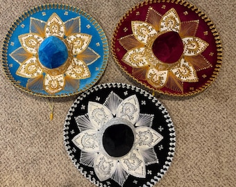 May include: Three wide-brimmed mariachi hats in blue, burgundy, and black. Each hat features intricate gold or silver metallic embroidery in a floral petal pattern radiating from the center crown, with decorative braided trim along the outer edge.