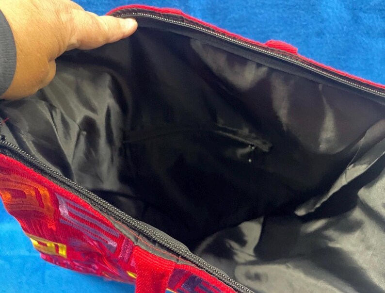 May include: A large tote bag featuring a vibrant red exterior with a geometric pattern in shades of yellow and purple. The bag has a black zippered closure at the top and a black interior lining with a small zippered pocket on the inside wall.