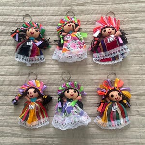 May include: Six fabric doll keychains featuring embroidered faces, dark hair, and colorful ribbon headpieces. Each doll wears a patterned dress with lace trim and a metal keyring. The dolls are labeled with the numbers 25, 26, 27, 28, 29, and 30.
