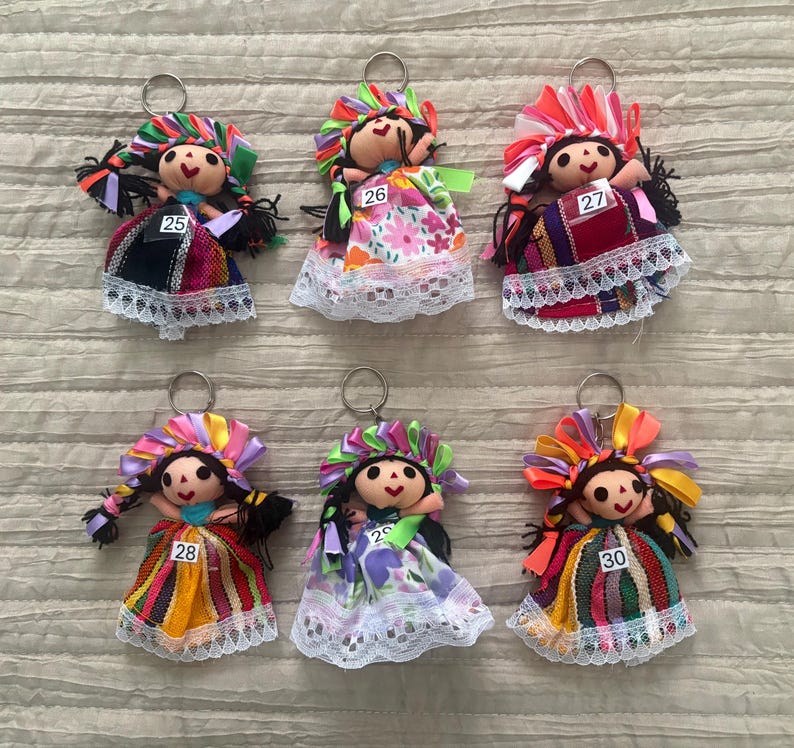May include: Six fabric doll keychains featuring embroidered faces, dark hair, and colorful ribbon headpieces. Each doll wears a patterned dress with lace trim and a metal keyring. The dolls are labeled with the numbers 25, 26, 27, 28, 29, and 30.
