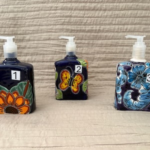 Puede incluir: Tres dispensadores de jabón de cerámica azul oscuro con bombas blancas. Presentan diseños pintados a mano: una flor, una mariposa y motivos florales azules. Cada dispensador está numerado con un cuadrado blanco.