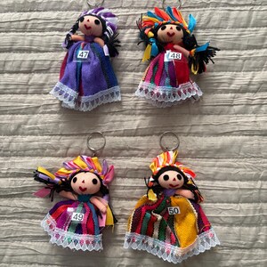 May include: Four handmade doll keychains featuring black yarn hair, embroidered facial features, and colorful fabric dresses with white lace trim. Each doll wears a headpiece made of ribbons and has a white tag numbered 47, 48, 49, or 50.