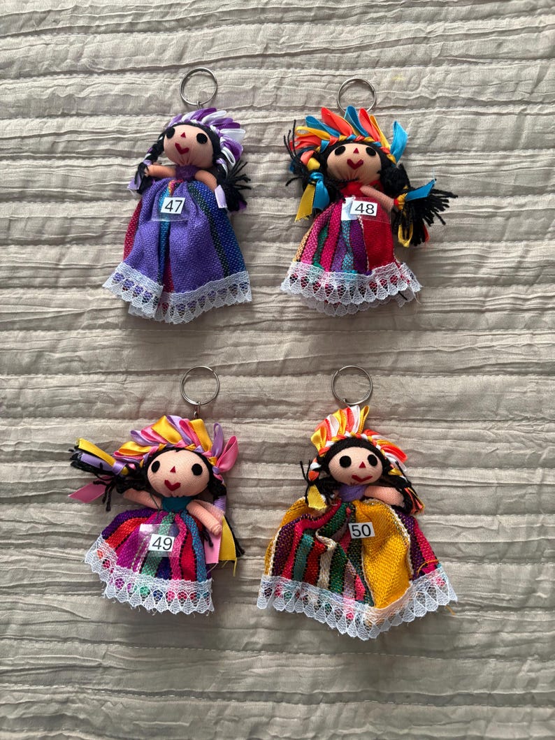 May include: Four handmade doll keychains featuring black yarn hair, embroidered facial features, and colorful fabric dresses with white lace trim. Each doll wears a headpiece made of ribbons and has a white tag numbered 47, 48, 49, or 50.