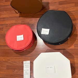 May include: Three round hat cases in camel, black, and salmon, and one white octagonal hat box. Each item has a white label identifying it as HAT CASE CAMEL, HAT CASE BLACK, HAT CASE SALMON, or HAT BOX. A small white note states THESE ITEMS ARE IN A LISTING: HAT CASE AND HAT BOX.