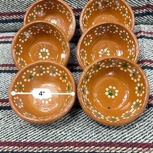 Puede incluir: Seis cuencos pequeños de terracota pintados a mano, cada uno de aproximadamente 10 cm de diámetro, están dispuestos sobre un textil a rayas. Los cuencos son de color marrón cálido con patrones florales blancos y verdes alrededor de los bordes y centros. El fondo es una tela texturizada.