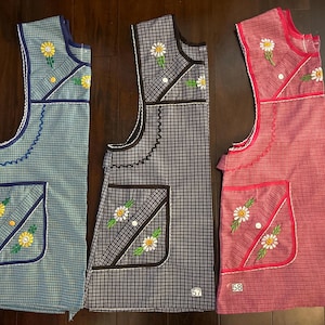 May include: Three vintage aprons in blue, grey, and red, each with a gingham pattern and daisy embroidery. The aprons have pockets and decorative trim. Numbers 56, 57, and 58 are visible on the aprons.