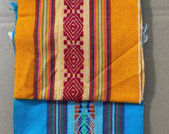 May include: Three colorful woven textiles stacked on top of each other. The top textile is orange with a central red and cream diamond pattern. The middle textile is blue with a central geometric design. The bottom textile is pink with a central geometric design.