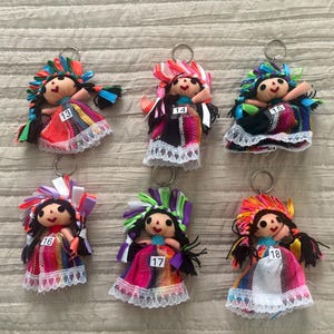 May include: Six fabric doll keychains with black yarn hair, colorful ribbon headpieces, and dresses featuring woven patterns and white lace trim. Each doll has a small white tag numbered 13, 14, 15, 16, 17, or 18 attached to the front of the dress.