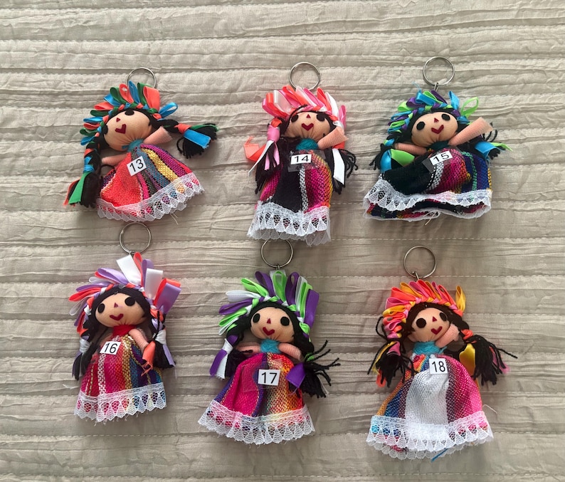 May include: Six fabric doll keychains with black yarn hair, colorful ribbon headpieces, and dresses featuring woven patterns and white lace trim. Each doll has a small white tag numbered 13, 14, 15, 16, 17, or 18 attached to the front of the dress.