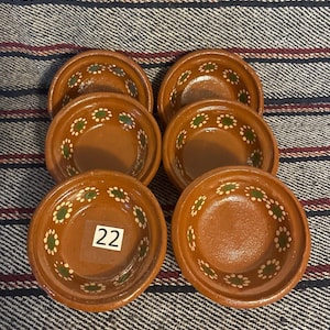 Peut inclure: Un ensemble de six petits bols ronds en terre cuite brune disposés en deux colonnes. Chaque bol présente un bord intérieur peint à la main avec un motif répétitif de points blancs et verts en forme de fleurs. Un bol porte une étiquette carrée blanche avec le numéro 22.