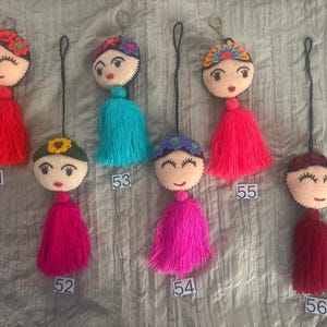 May include: Six felt doll head keychains with embroidered facial features and colorful yarn tassels. Each doll has a unique hair accessory, including flowers and headbands. The keychains are labeled with numbers 51 through 56 for identification.