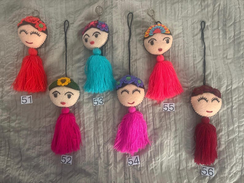 May include: Six felt doll head keychains with embroidered facial features and colorful yarn tassels. Each doll has a unique hair accessory, including flowers and headbands. The keychains are labeled with numbers 51 through 56 for identification.