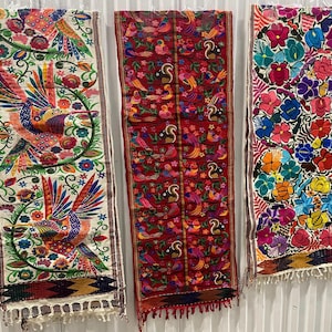 Peut inclure: Trois chemins de table brodés colorés avec des franges. Le chemin de table de gauche a un fond blanc avec des oiseaux multicolores et des motifs floraux. Le chemin de table du milieu a un fond rouge avec des oiseaux multicolores et des motifs floraux. Le chemin de table de droite a un fond blanc avec des motifs floraux multicolores.