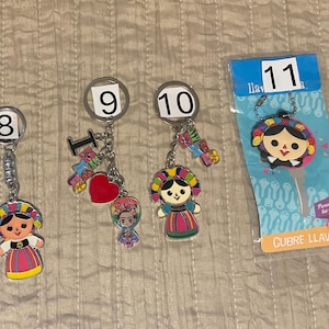 Puede incluir: Cinco llaveros coloridos con un diseño tradicional de muñeca mexicana. Cada llavero tiene un anillo de plata y una variedad de dijes, incluyendo corazones, flores y la palabra "Me encanta". Los llaveros están etiquetados con los números 8, 9, 10 y 11.