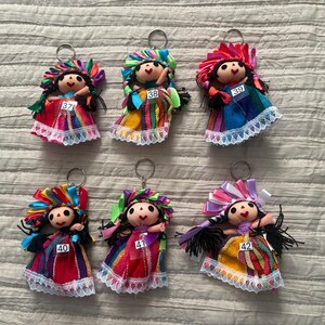 May include: Six fabric doll keychains with black yarn hair, colorful ribbon headpieces, and dresses featuring striped patterns and white lace trim. Each doll is labeled with a white tag numbered 37, 38, 39, 40, 41, or 42 attached to the front.