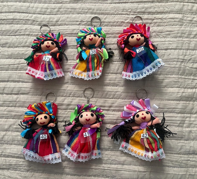 May include: Six fabric doll keychains with black yarn hair, colorful ribbon headpieces, and dresses featuring striped patterns and white lace trim. Each doll is labeled with a white tag numbered 37, 38, 39, 40, 41, or 42 attached to the front.