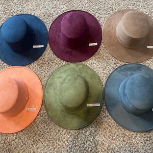 May include: Six wide-brimmed hats in various colors, including blue, purple, tan, orange, green, and teal. Each hat has a cylindrical crown and a flat brim. Small labels with color names are visible on each hat.